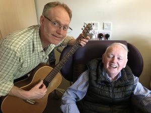Man with guitar and man in chair smiling