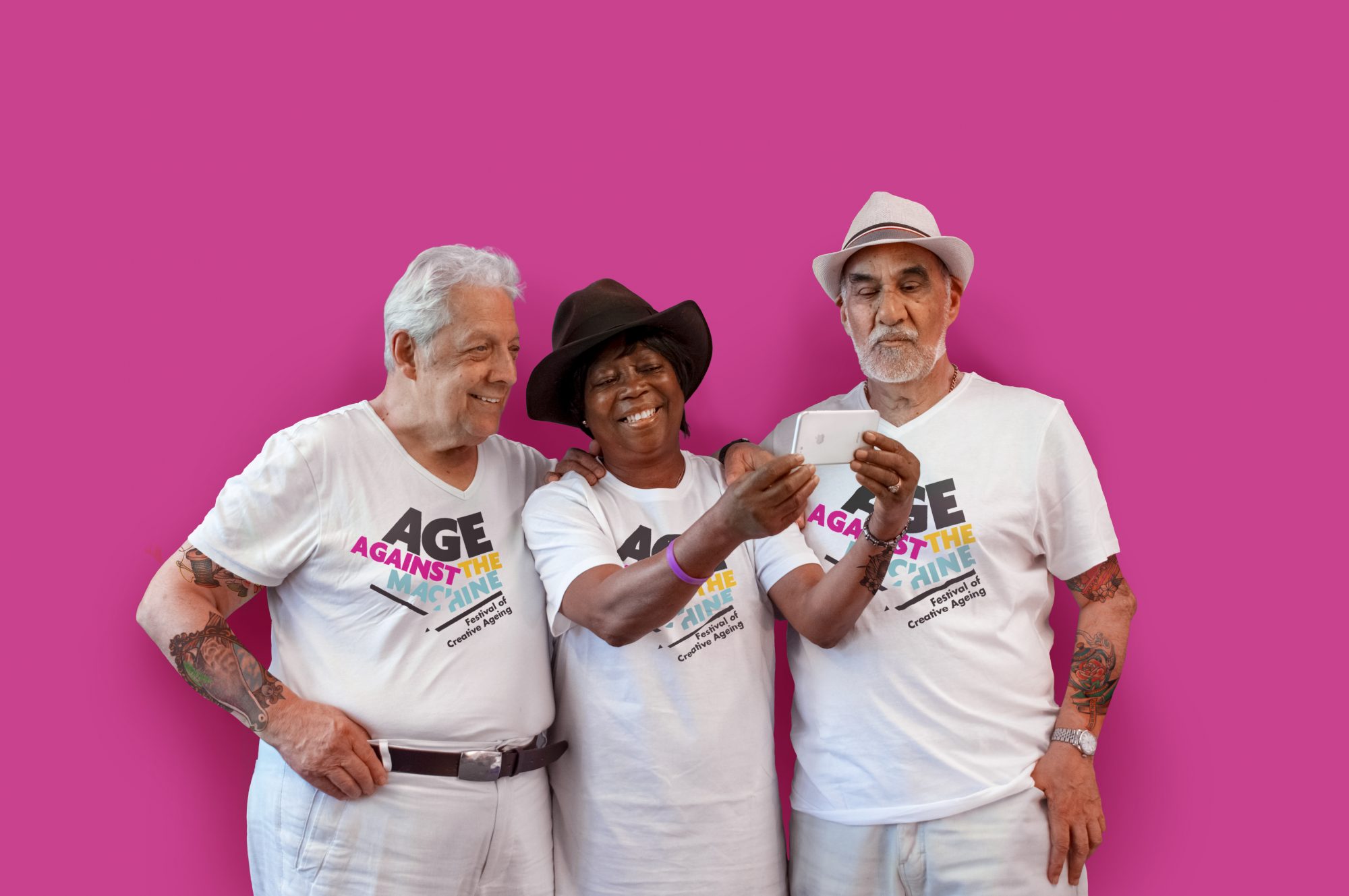 Three smiling people standing in front of a pink wall looking at something on an iphone.