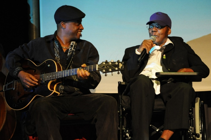 man in wheelchair with microphone and man with guitar smiling