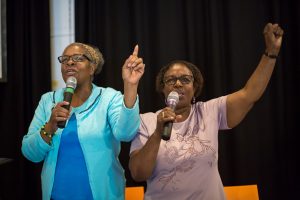 two elder women singing with microphones arms raised smiling 