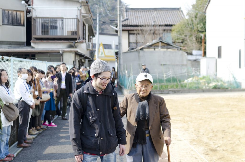 one older and one younger japanese men walking along a path with people gathered in a line watching them.