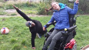 White woman in a wheelchair and a black man on the grass reaching up they are smiling