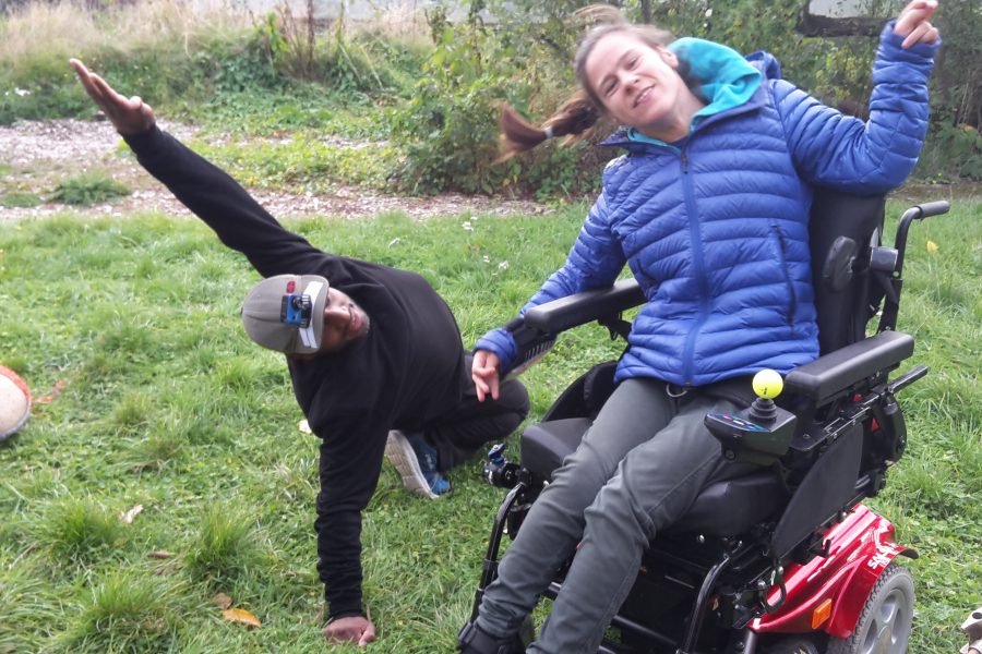 White woman in a wheelchair and a black man on the grass reaching up they are smiling