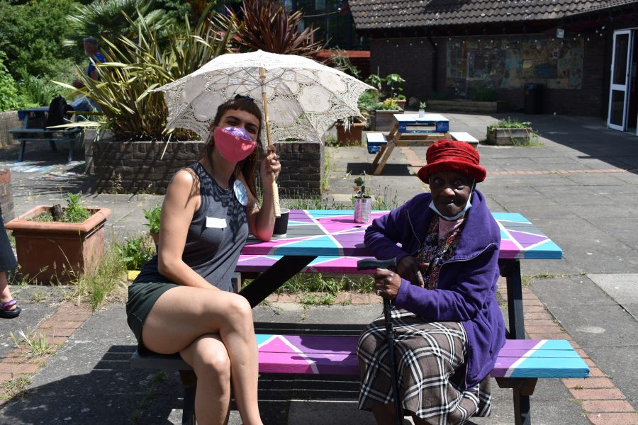 Photo of a young white woman and an older black woman sitting on a bench outside on a sunny day.