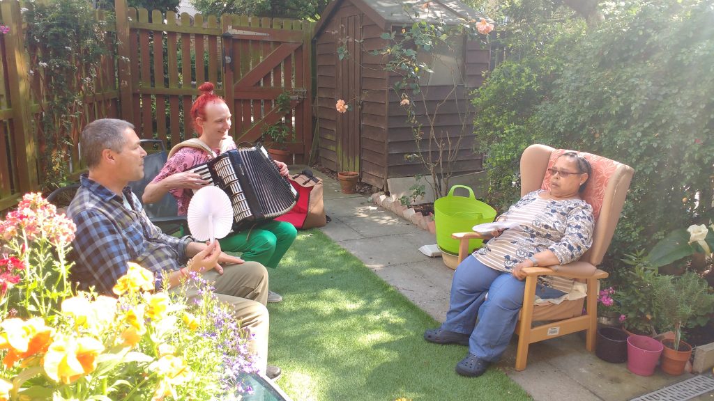 Two men and a woman sitting in a garden. One of the man is playing the accordion.