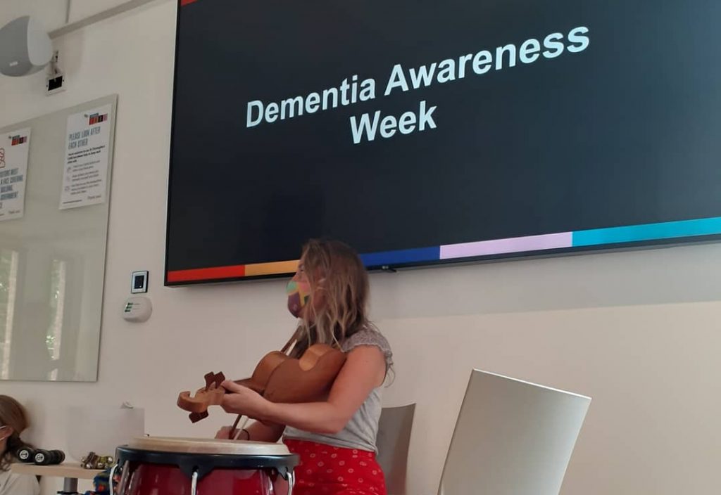 A blonde woman plays the violin. Behind her a screen read Dementia Awareness Week.