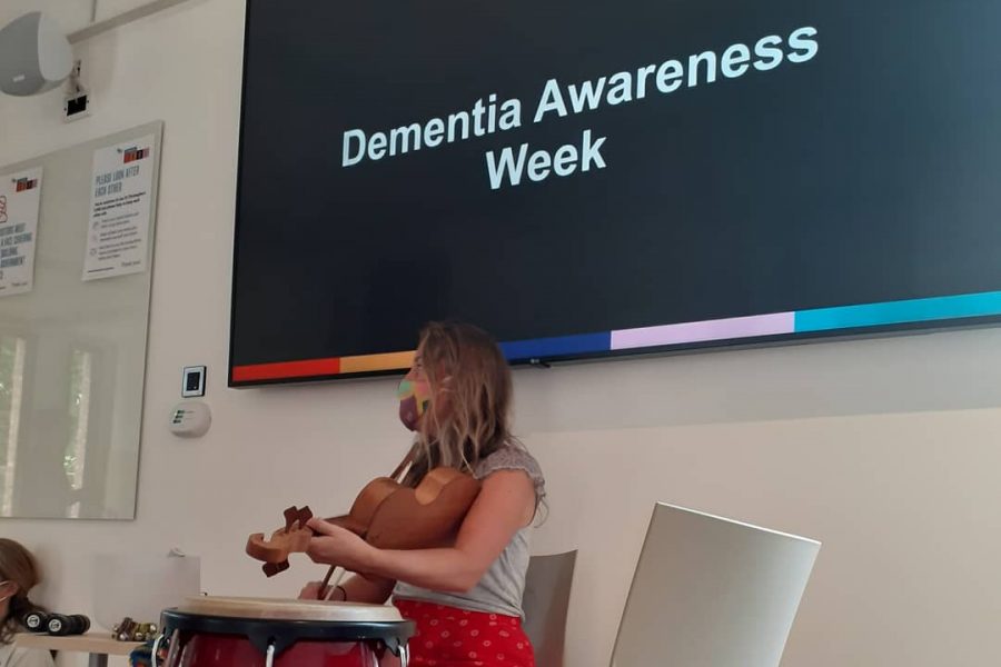 A blonde woman plays the violin. Behind her a screen read Dementia Awareness Week.
