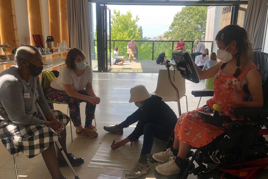 A black older woman and a white woman sit on chairs looking at the woman in front of them on a wheelchair using a text to speech keyboard. Another woman sits on the floor taking notes on a piece of paper. They're all wearing masks.