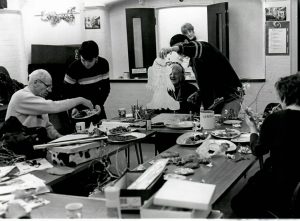 Black and white photo of a group of older people sitting a tables full of arts and craft material. Two volunteers help them handling the material.