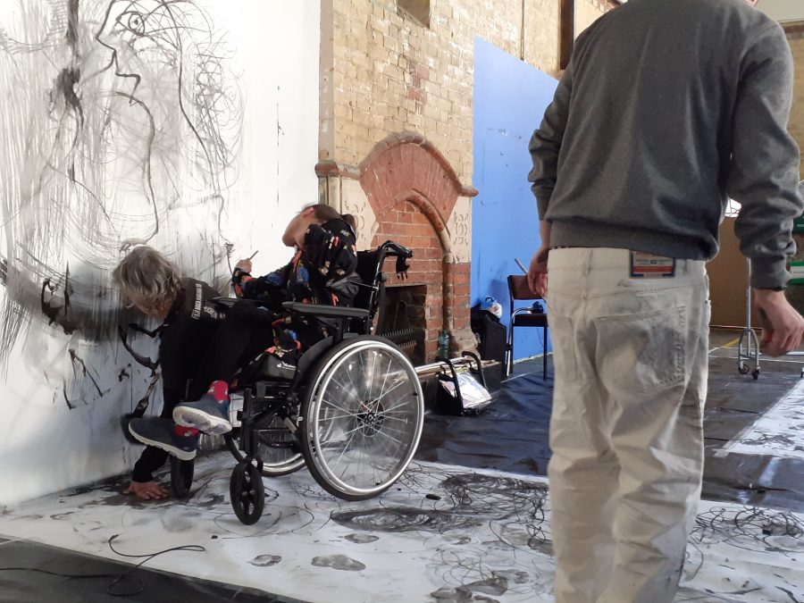A person in a wheelchair drawing with charcoal on a large piece of paper that is next to them on the wall and beneath them on the floor,