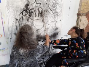 Two women make marks on a large sheet of paper on the wall. One of the women is in a wheelchair.