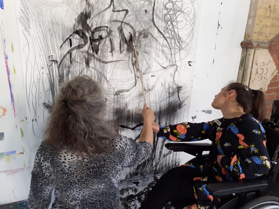 Two women make marks on a large sheet of paper on the wall. One of the women is in a wheelchair.