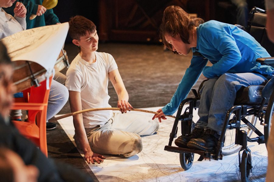 A person in a white T-shirt sits on a large piece of paper with a woman in a wheelchair. They draw together