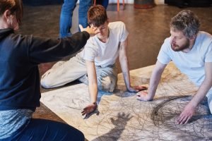 Two people in white T-shirts sitting on a large piece of paper that is covered in grey scribbles.