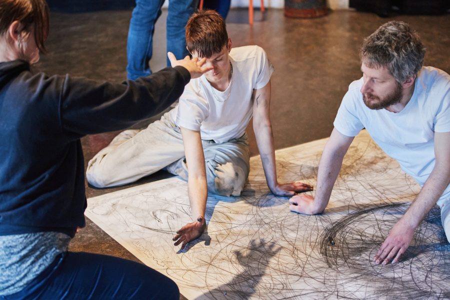Two people in white T-shirts sitting on a large piece of paper that is covered in grey scribbles.