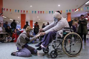 Two women wearing masks dance together. One woman is kneeling in front of an older woman, who is in a wheelchair. They are holding hands.