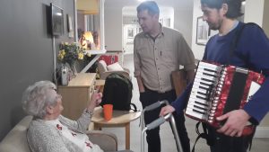 Two men stand in a corridor opposite an older woman who is sitting down. They are smiling. One of the men is holding an accordion.