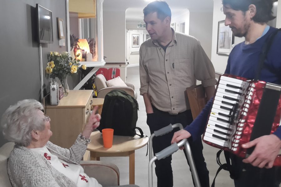 Two men stand in a corridor opposite an older woman who is sitting down. They are smiling. One of the men is holding an accordion.